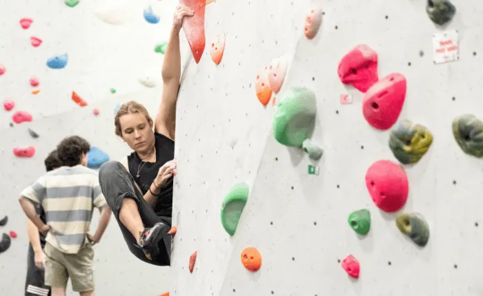 A young person in dark clothing climbing an indoor wall with coloured handholds whilst a child in a striped shirt watches from below.