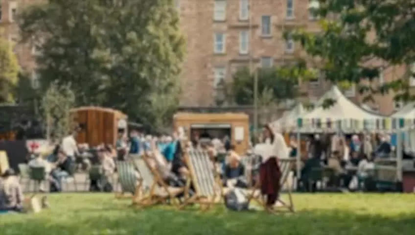 People relaxing in deckchairs on a sunny lawn with brick buildings and market stalls behind.