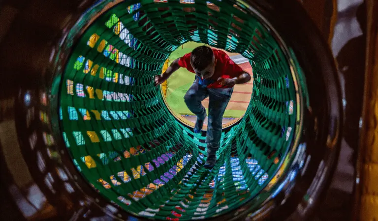 Child in red shirt crawling through a green mesh tunnel at an indoor play facility.