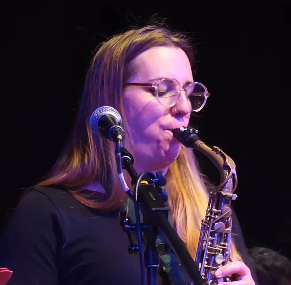 Musician performing on stage, singing into a microphone whilst playing saxophone, wearing glasses and a dark top under purple stage lighting.