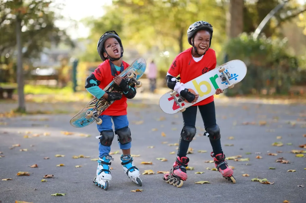 Two young skateboarders wearing helmets and protective gear hold their boards while rollerblading on a tree-lined path in autumn.