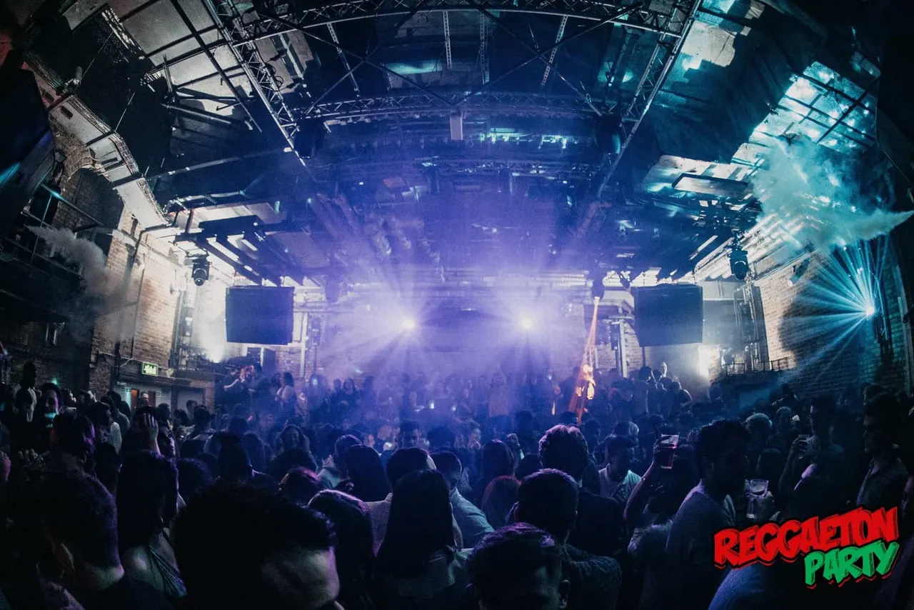 Large crowd dancing under dramatic blue and purple stage lighting in an indoor concert venue with industrial ceiling structure.