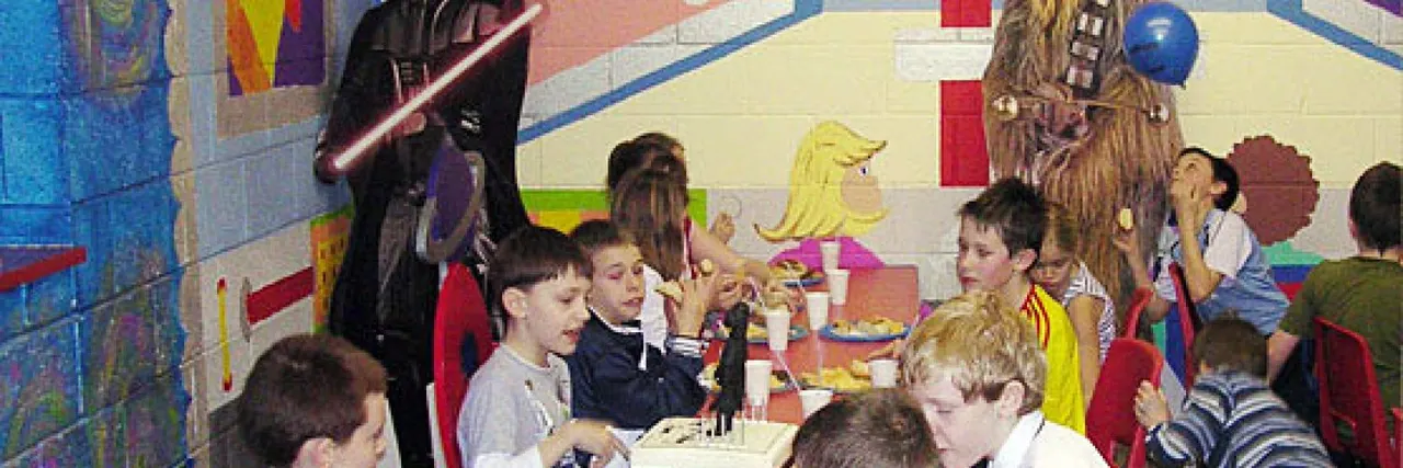 Children eating at a table during a themed party in a colourful indoor play venue with character decorations on the walls.