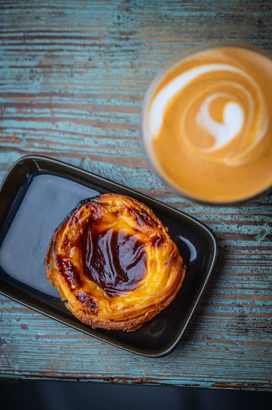 Pastry with chocolate drizzle on a black plate next to a latte with latte art on weathered blue wood.