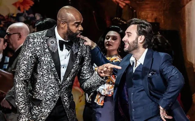 Guests in formal evening wear socialising at an indoor event, with one man in a patterned jacket conversing with another in a blue suit.