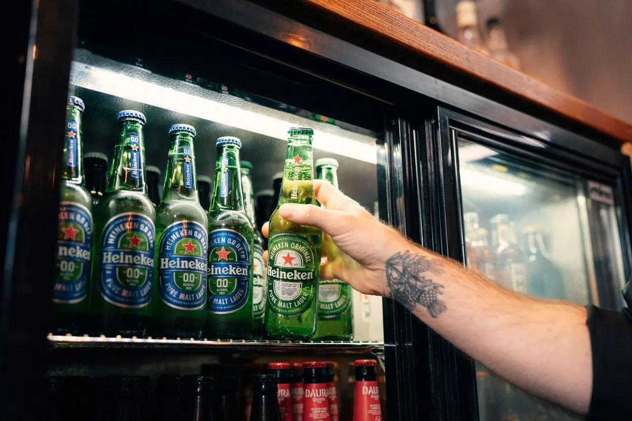 Person removing a beer bottle from an illuminated refrigerator display filled with green glass bottles.