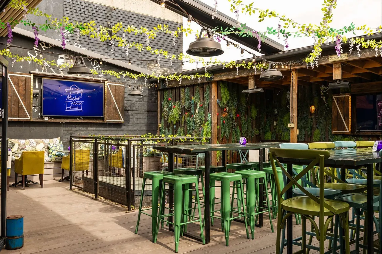 Outdoor rooftop terrace with green bar stools, dining tables, hanging foliage in yellow and purple, brick walls, and a neon sign.