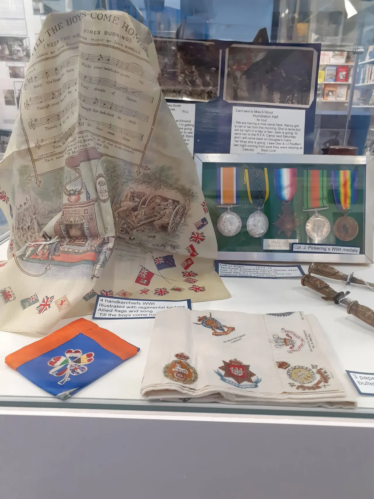 Museum display case with WWI memorabilia including sheet music, medals, handkerchiefs, badges and postcards from the First World War period.
