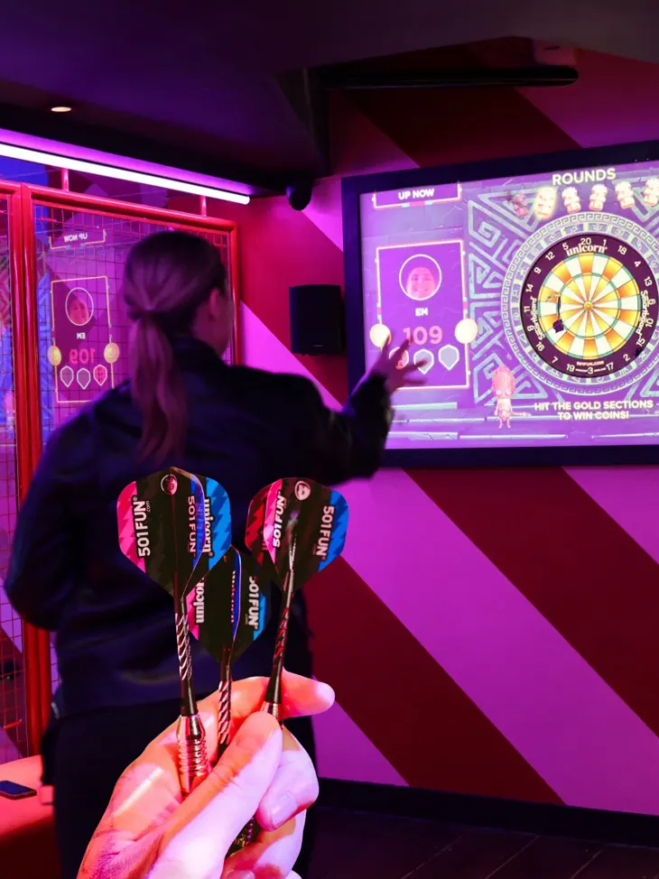 Person playing darts at an arcade bar with neon-lit gaming screens and colourful lighting.