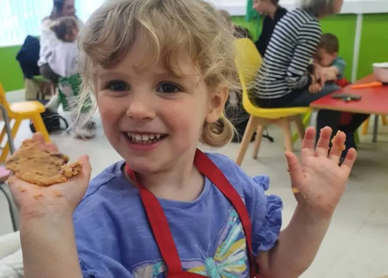 Young child holding a biscuit and waving at a children's activity centre with other children and adults in background.