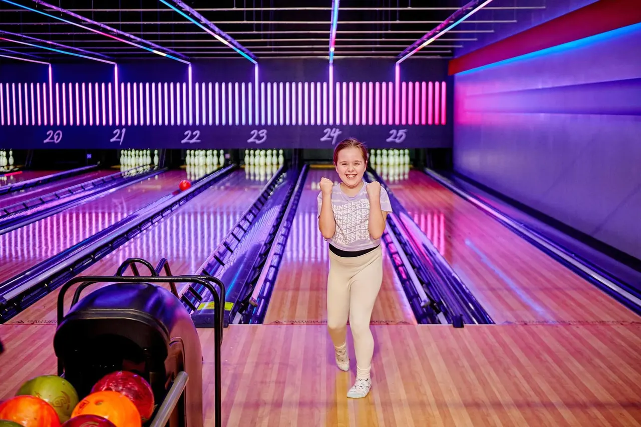Child celebrates with clenched fists on a neon-lit bowling lane surrounded by numbered lanes and coloured pins.
