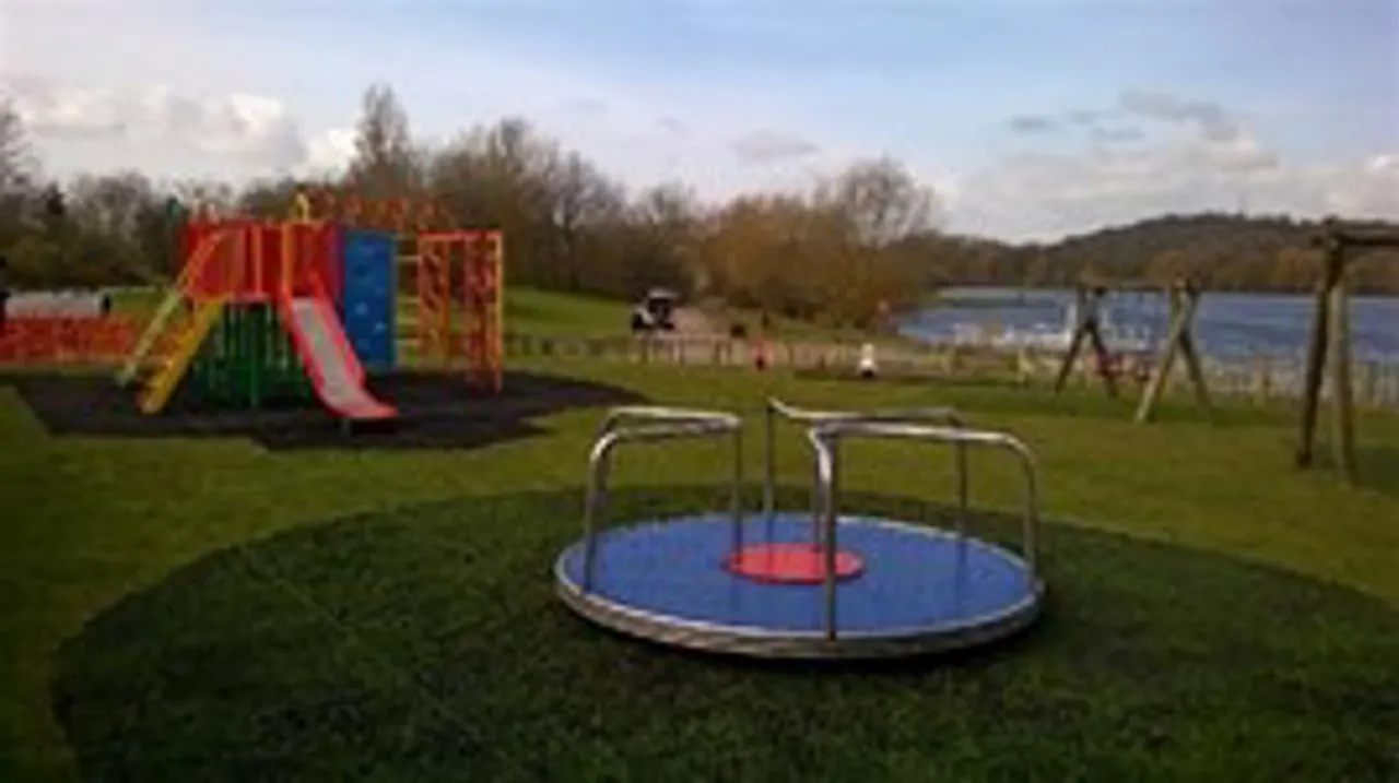 Outdoor play area with a low trampoline in foreground, colourful climbing frame and slide beyond, set in grassy parkland with water visible in distance.