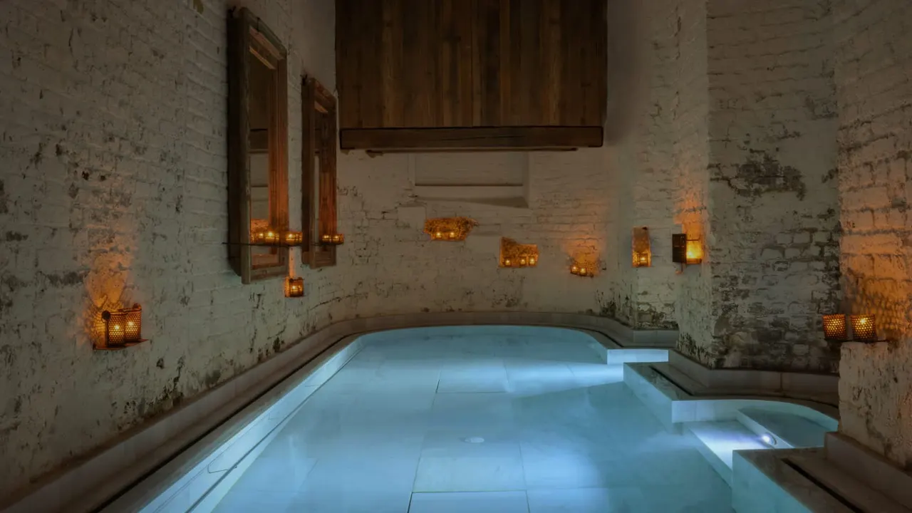 Underground spa pool lit by candlelight, with weathered brick walls and wooden fixtures reflected in still blue water.