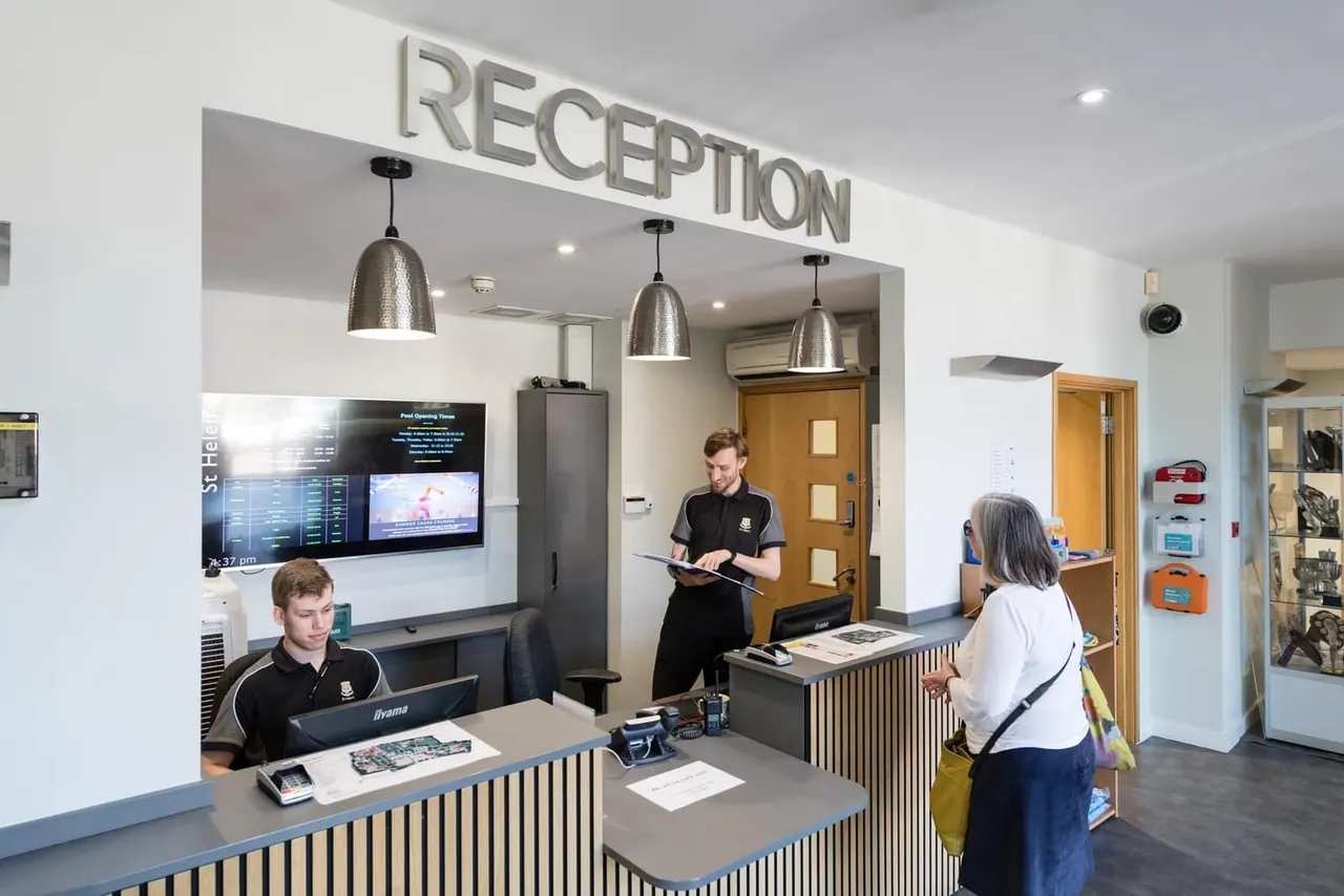 Reception desk with staff assisting visitors, modern white and wooden interior with pendant lights and digital displays.