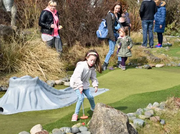 Girl playing mini golf on a course with families standing nearby watching.