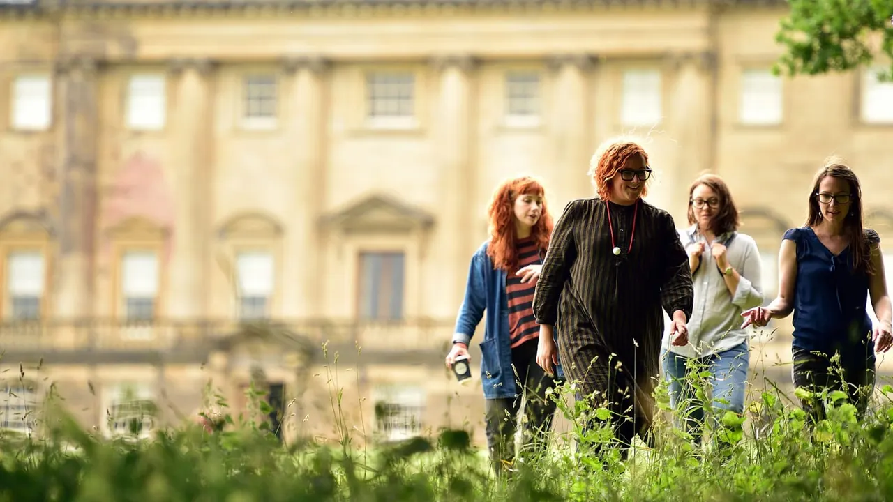 Four women walking through a wildflower meadow in front of a grand stone mansion.