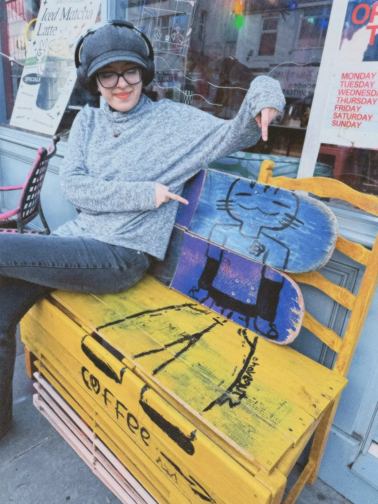 Young person wearing headphones and glasses posing with colourful skateboards outside a café window.