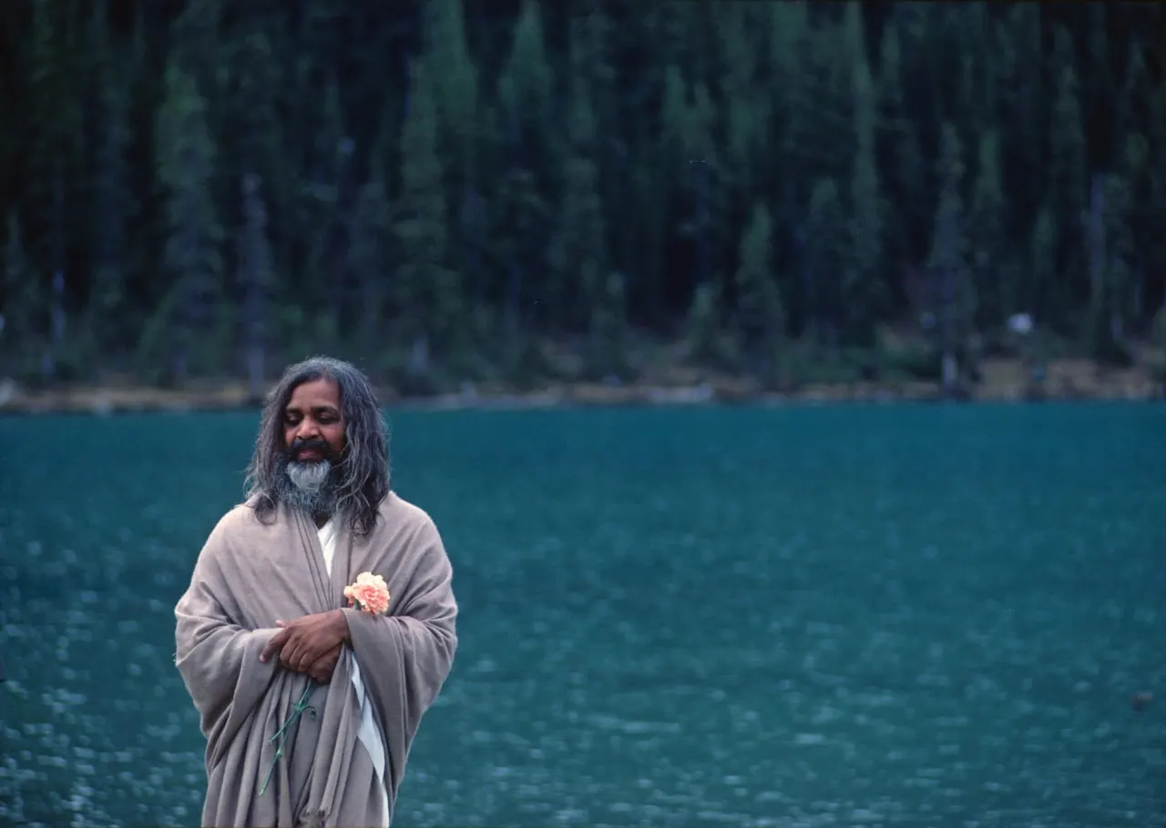 Man with long grey hair and beard holding a flower, standing by a turquoise lake with evergreen forest behind.
