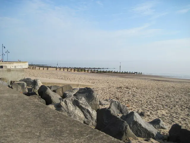 Sandy beach with a stone breakwater and pier extending into calm sea under blue sky.