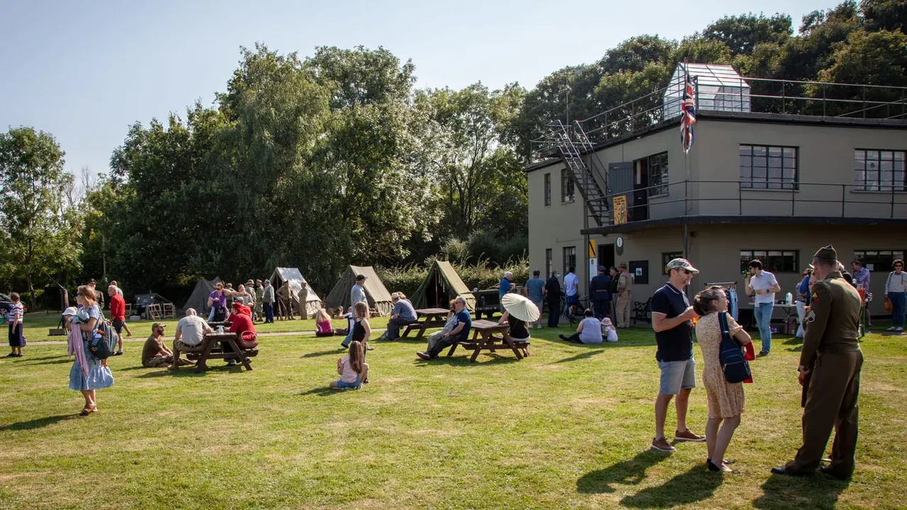 Visitors in period costume and modern dress gather on a sunny grass field with canvas tents, picnic tables, and a two-storey control tower building flying the Union Jack.
