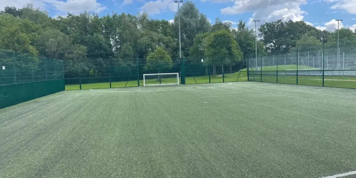 Empty artificial football pitch with goal, surrounded by green fencing and trees under blue sky.