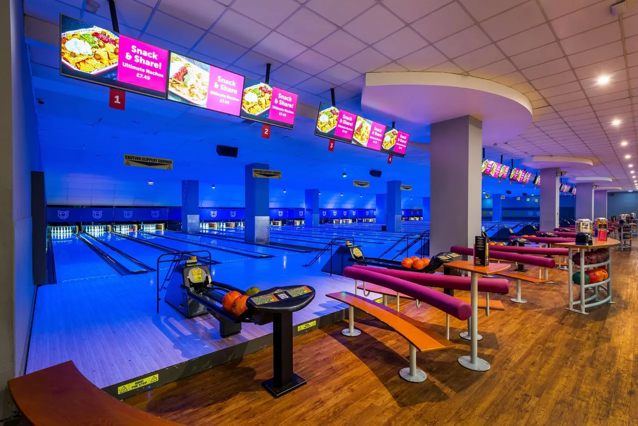 Interior of a bowling alley with multiple blue lanes, seating areas with burgundy benches, and illuminated menu boards advertising snacks and shakes.