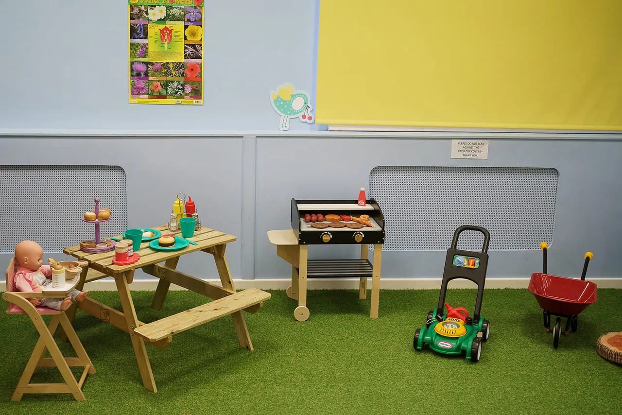 Play area with wooden picnic table, toy BBQ grill, toy lawn mower, pram and doll arranged on artificial grass.