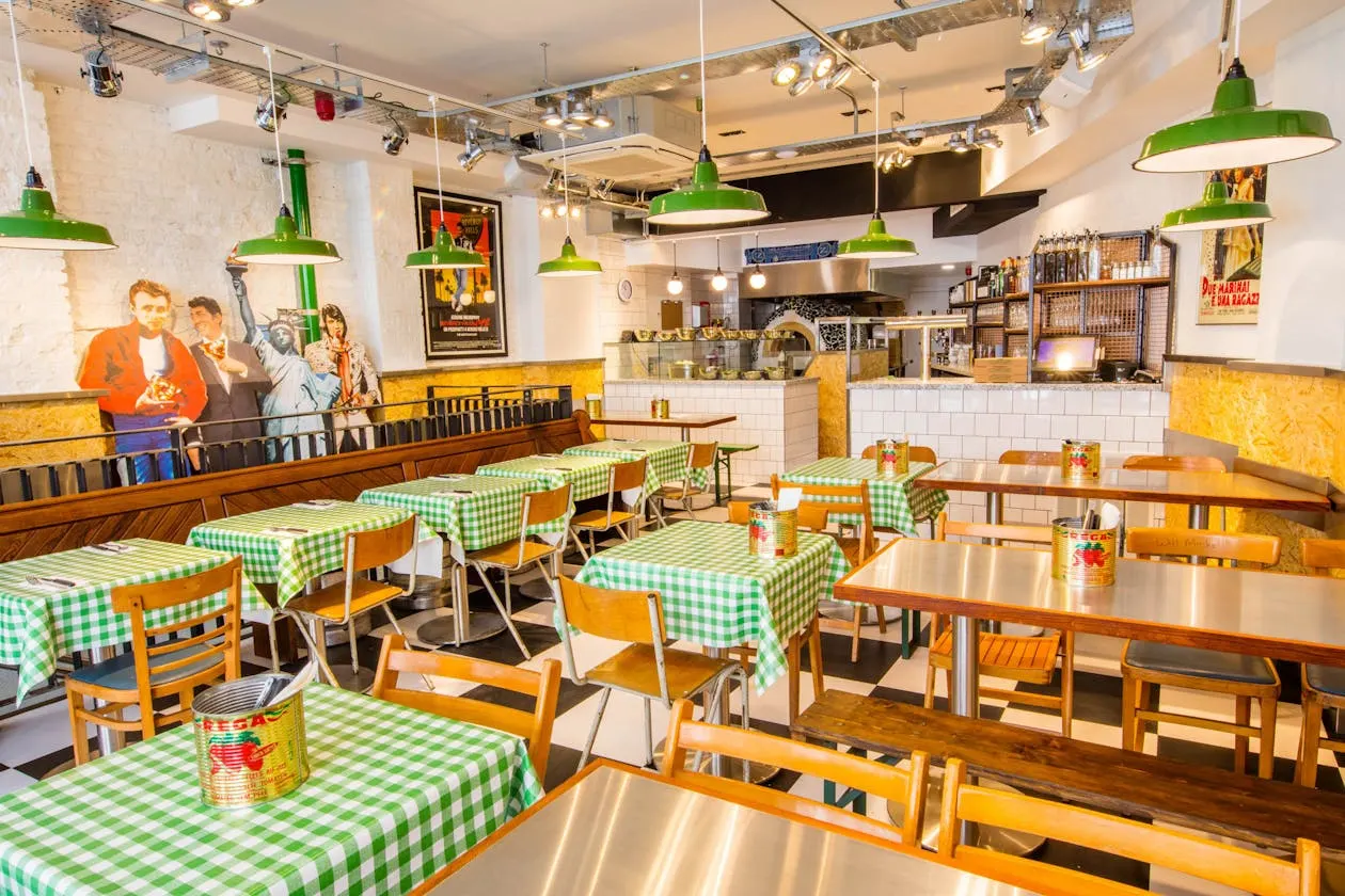 Retro-style pizzeria dining room with green-and-white checked tablecloths, wooden chairs, green pendant lights, and vintage film posters on white-tiled walls.