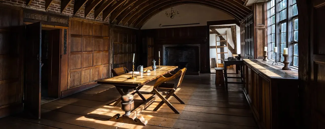 Historic timber-framed interior with long wooden dining table, period chairs, and arched ceiling lit by natural light from tall windows.