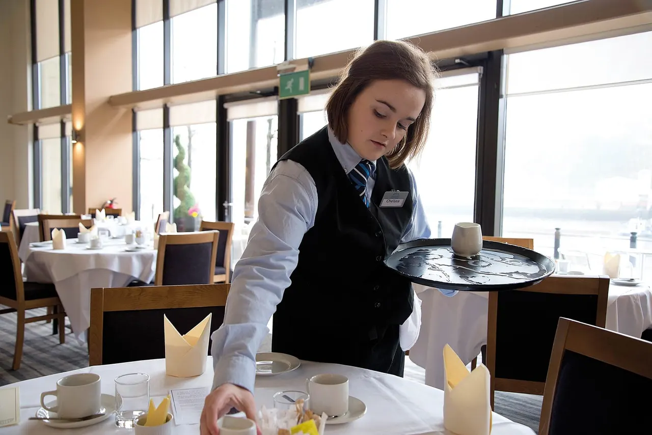 A waitress in uniform serves a dish at a fine dining table with a cityscape view visible through floor-to-ceiling windows.