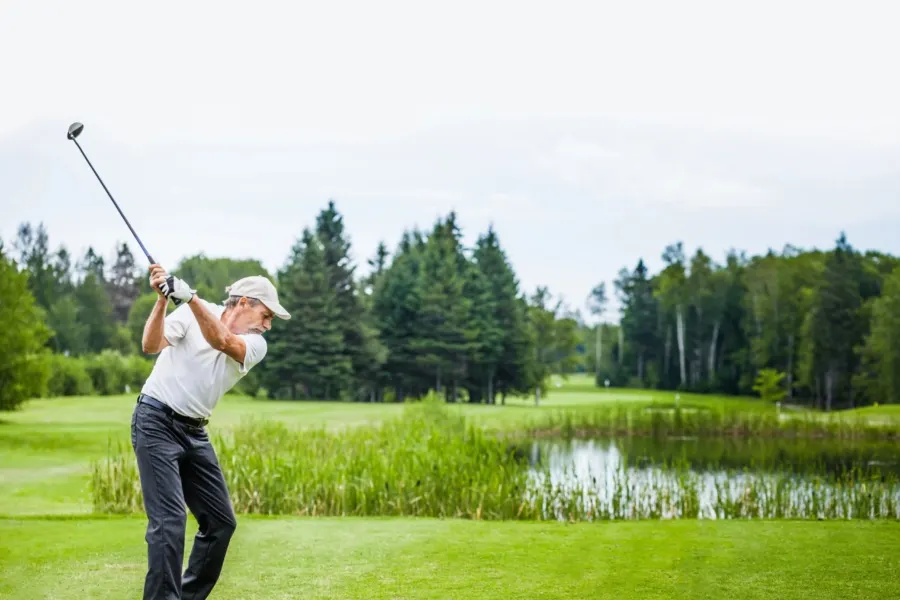 A golfer in white shirt and cap swings a golf club on a course with a pond, fairway and tree-lined background.