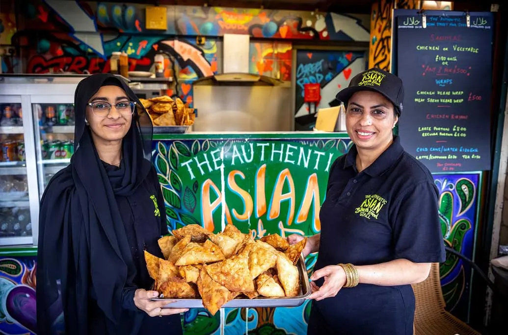 Two staff members hold a tray of fried samosas in a colourful Asian food restaurant interior.