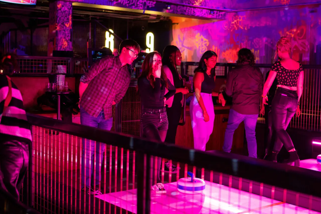 A group of people bowling and socialising under vibrant pink and purple neon lighting in a modern bowling alley.