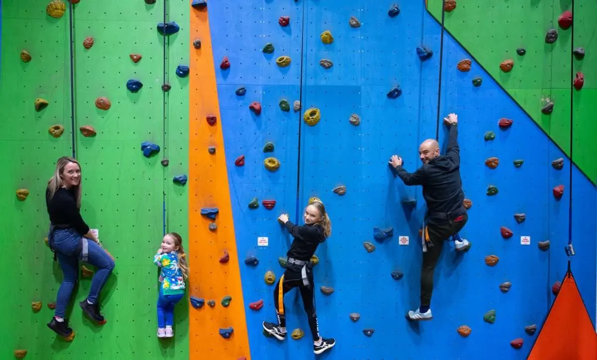 Four people of varying ages climbing an indoor rock wall with colorful holds on panels of green, blue and orange.
