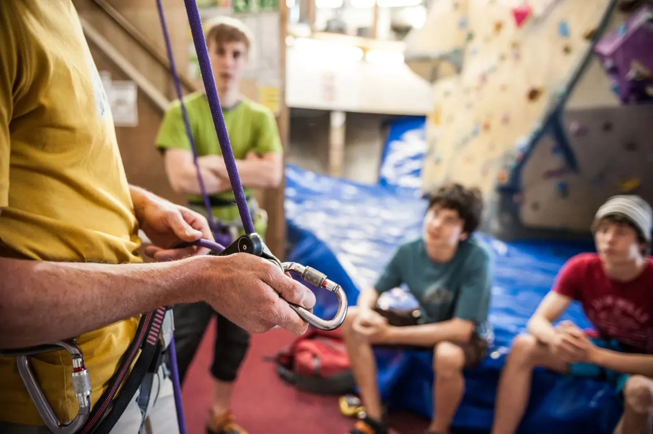 An instructor holds climbing equipment and safety rope whilst children watch in an indoor climbing wall facility.