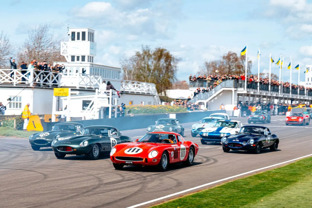 Classic racing cars competing on a circuit with spectators watching from grandstands and a white control tower building.