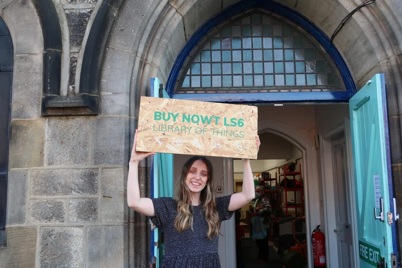 Young woman holding a wooden sign above her head at the entrance to a library of things, smiling at the camera.
