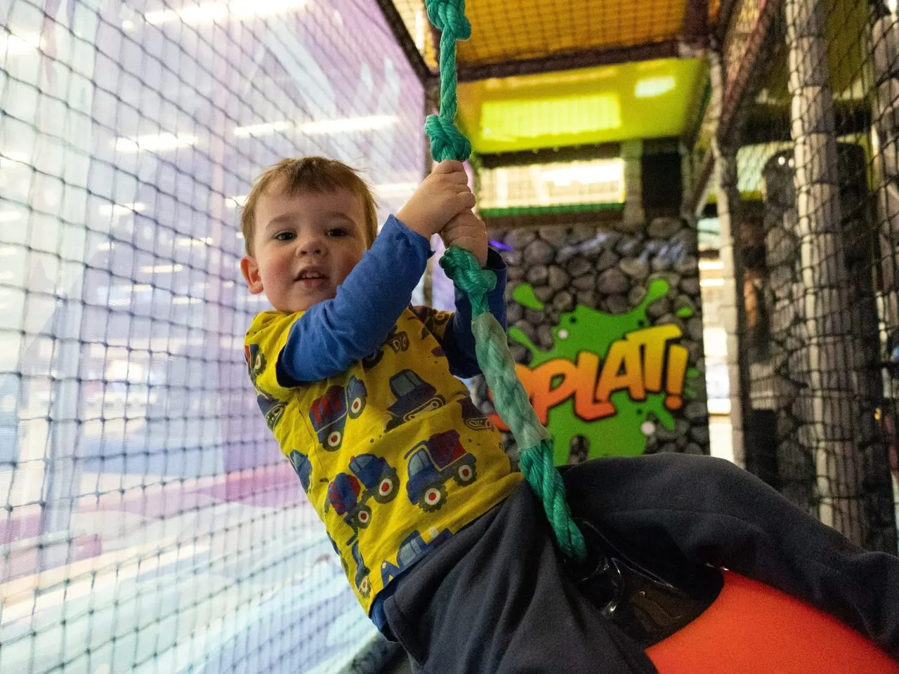 Young child climbing on a green rope in an indoor play area with coloured netting and signage.