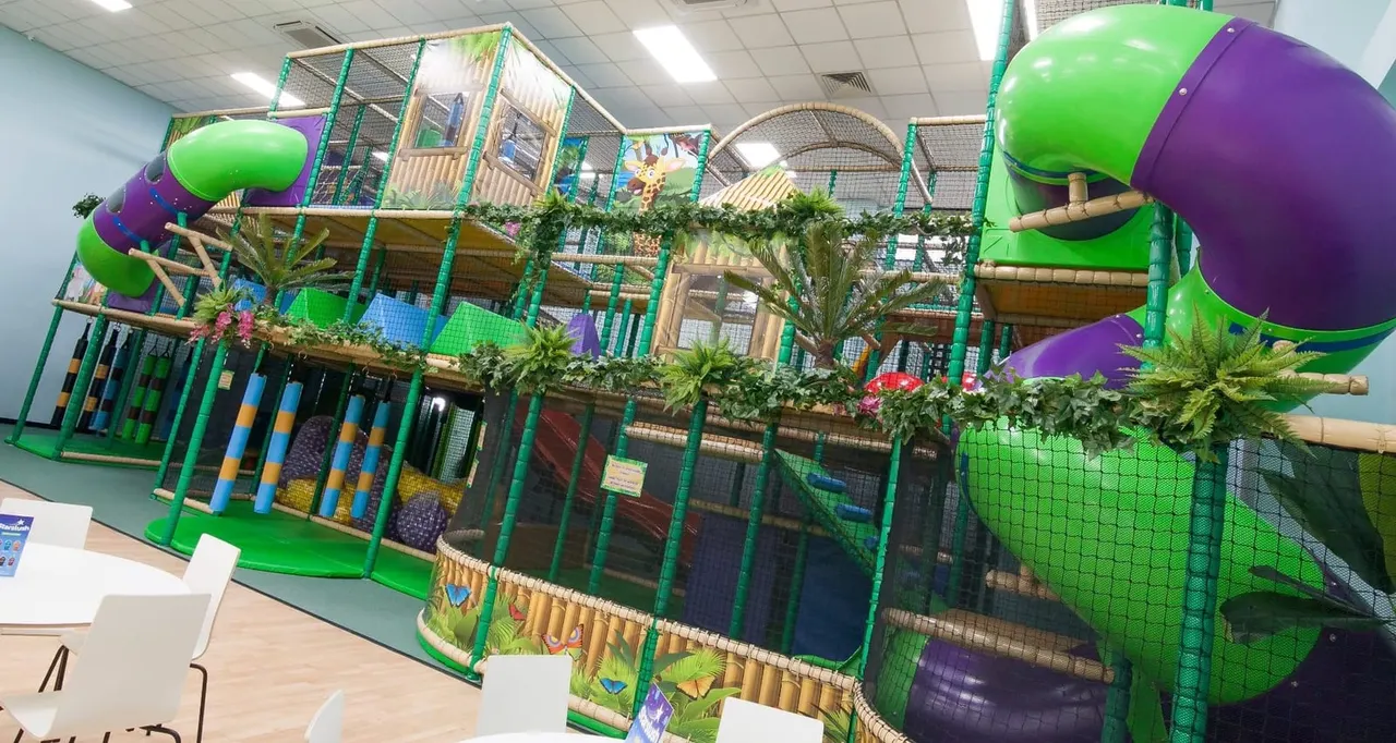 Large indoor play structure with green and purple tubes, slides, climbing nets and platforms decorated with plants and tropical themes.