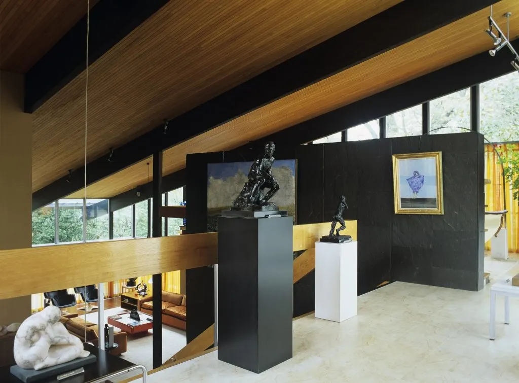 Gallery interior with sculptures on pedestals, framed artworks on black walls, wooden beams, and mezzanine level visible through glass railing.