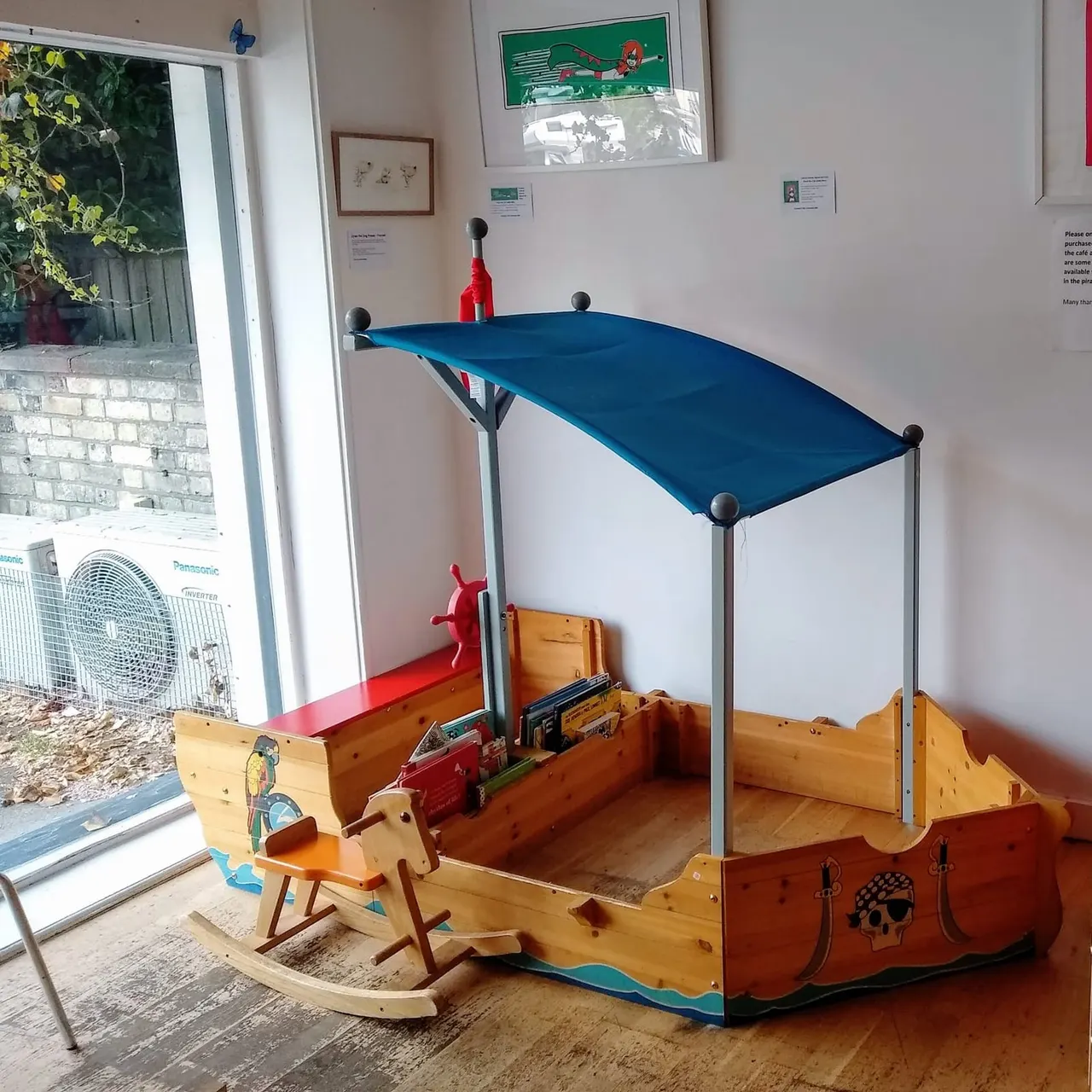 A wooden pirate ship-themed play structure with a blue canopy roof and rocking chair, set up indoors near glass doors opening to a garden.