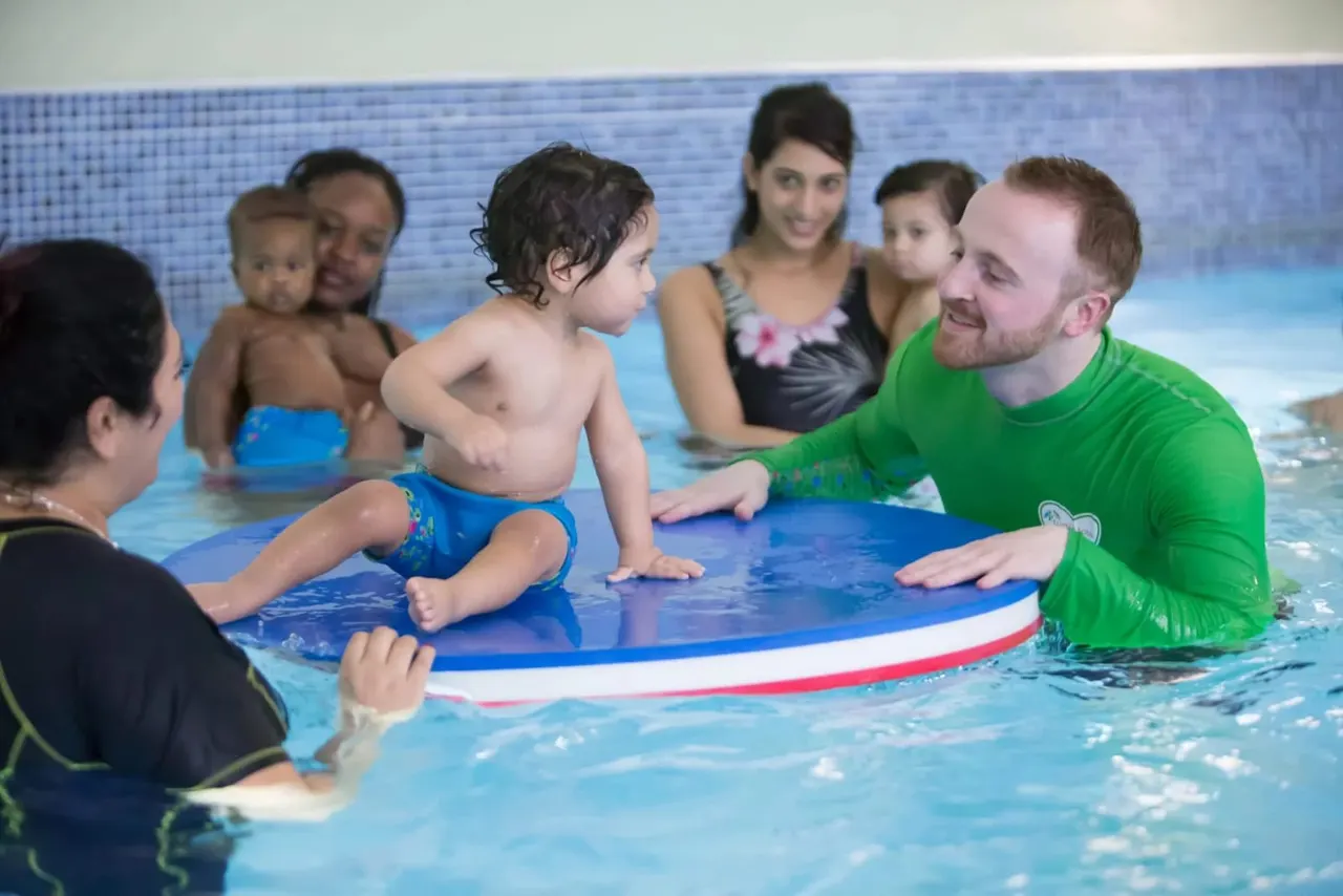 Instructor in green shirt teaching young children swimming skills on a blue float in indoor pool with parents watching.