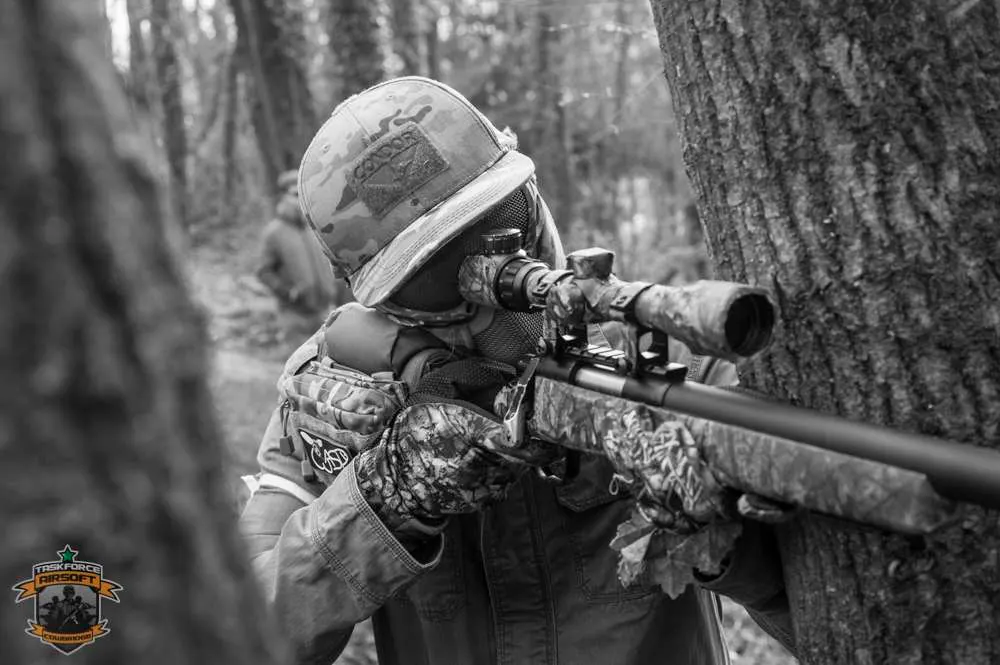Person in protective gear aiming a paintball marker rifle in a wooded paintball arena.