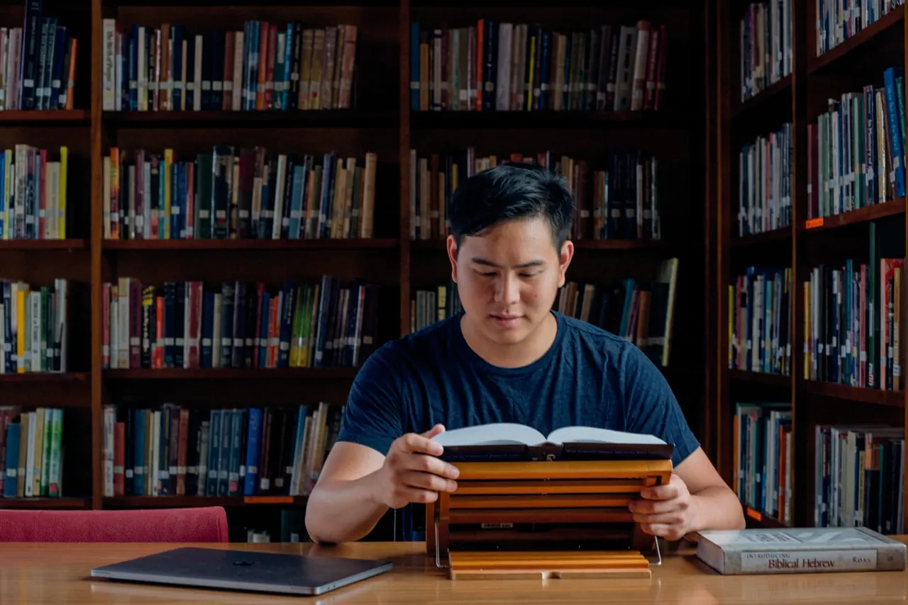A young man reads an open book at a library table with bookshelves behind him.