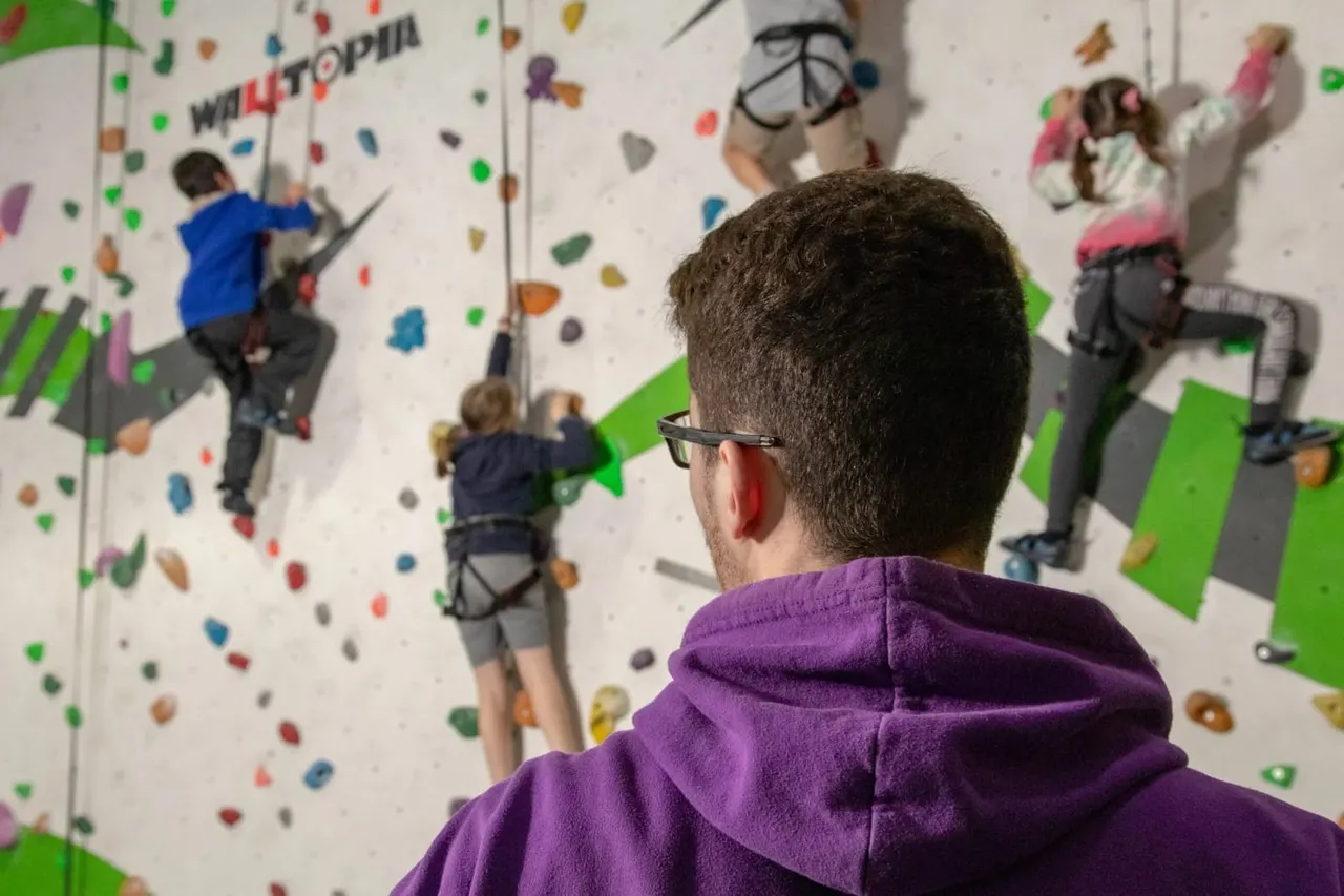An adult in a purple hoodie watches children climbing an indoor wall with coloured holds and safety ropes.