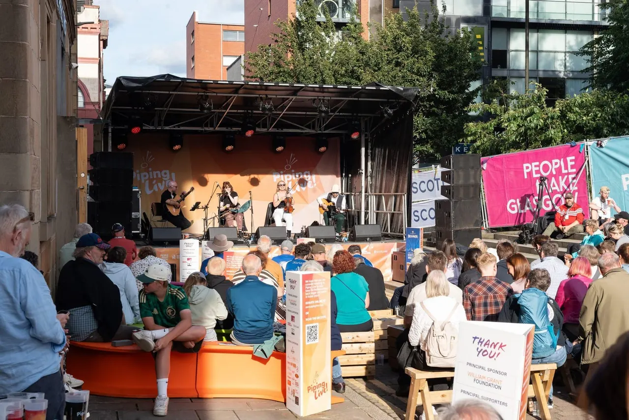 Outdoor concert with musicians performing on stage to a seated audience in an urban square surrounded by tall buildings.