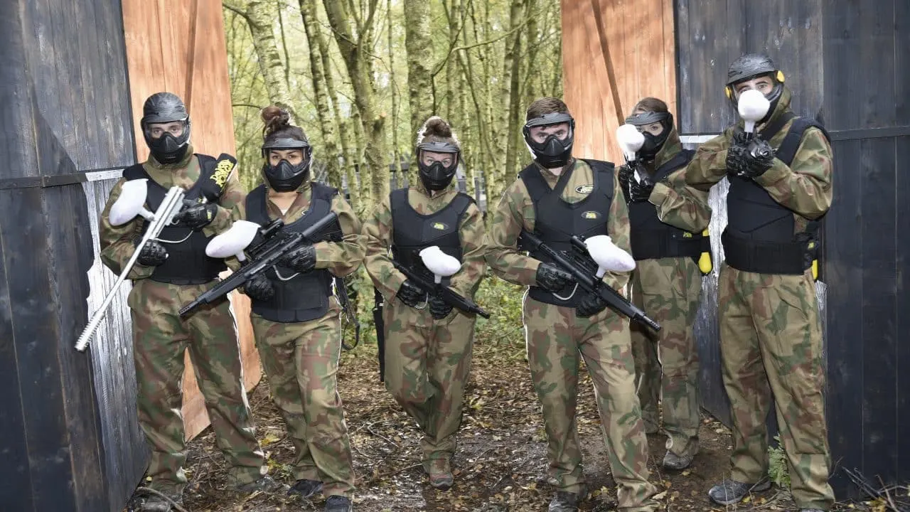Six paintball players in camouflage gear and protective masks holding paintball guns, posed in a wooded arena between wooden structures.