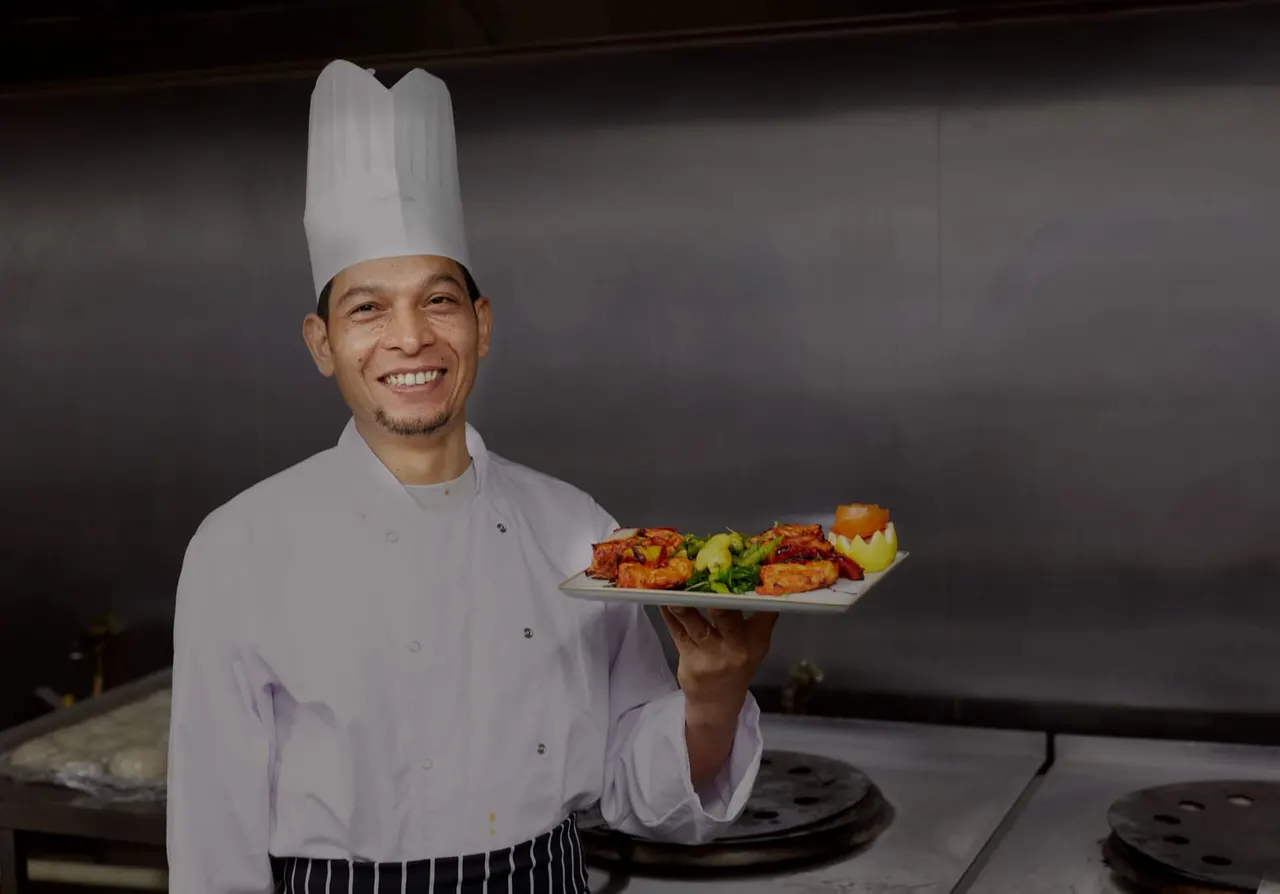 Chef in white uniform and tall hat holding a plate of cooked meat with vegetables and potatoes in a kitchen.