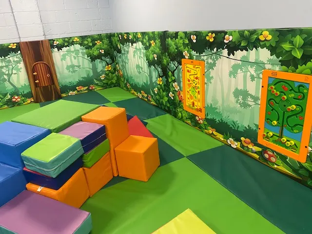 Indoor play area with colourful foam blocks and stepping platforms on a patchwork floor, with forest-themed wall graphics.