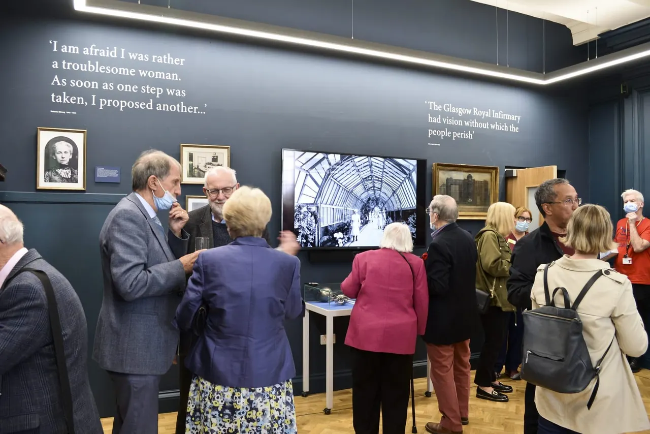 A museum guide leads a group of older visitors through an exhibition with dark blue walls displaying historical photographs and quotes about Glasgow Royal Infirmary.